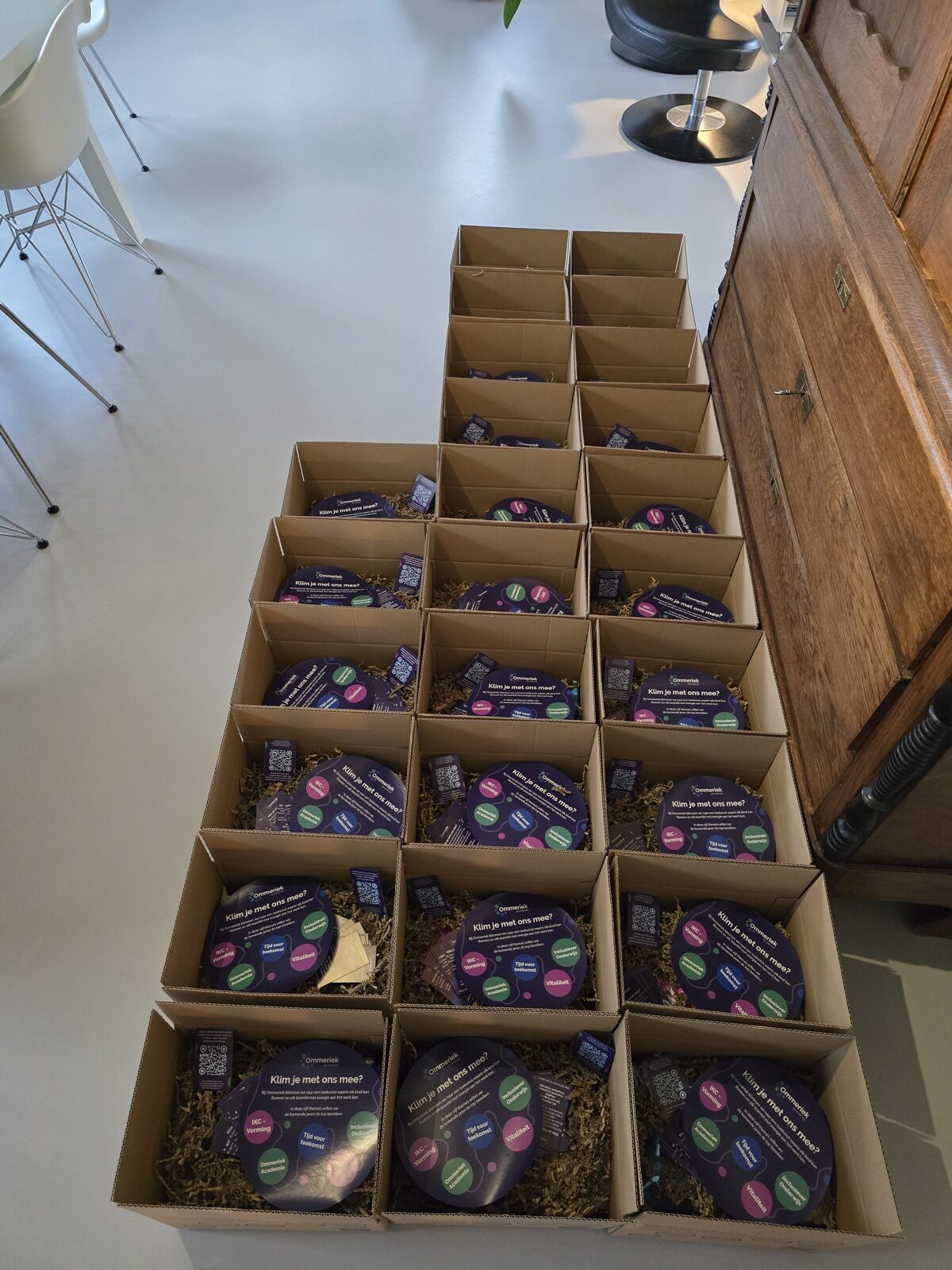 Vijfentwintig open vouwdozen met ronde flyers op een rij op de vloer, klaar om naar de scholen van Ommeriek te gaan